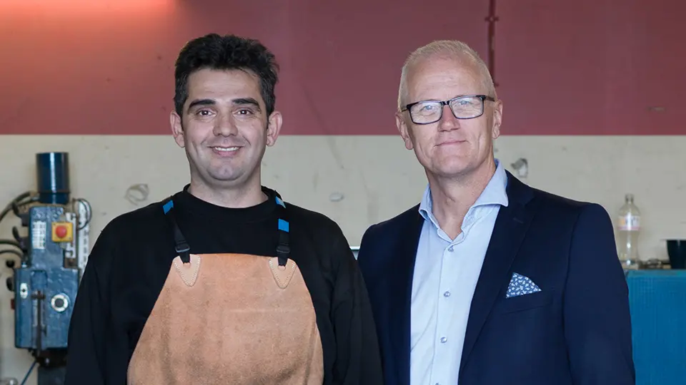 Två män i en verkstad. Till vänster en arbetare med skyddsförkläde, bredvid honom en man i kostym.